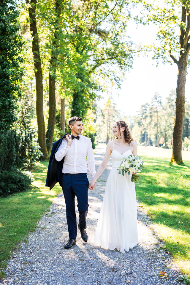 Brautpaar geht Hand in Hand beim Hochzeits-Shooting in Bielefeld. Der Bräutigam hat mit der anderen Hand sein Sakko über die Schulter geworfen. Beide schauen sich glücklich an und befinden sich inmitten grüner Natur.