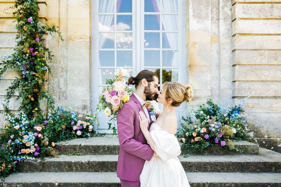 Brautpaar küsst sich vor seinem französischen Hochzeitsschloss. Üppige Blumendeko ist auf den Treppenstufen zu sehen.