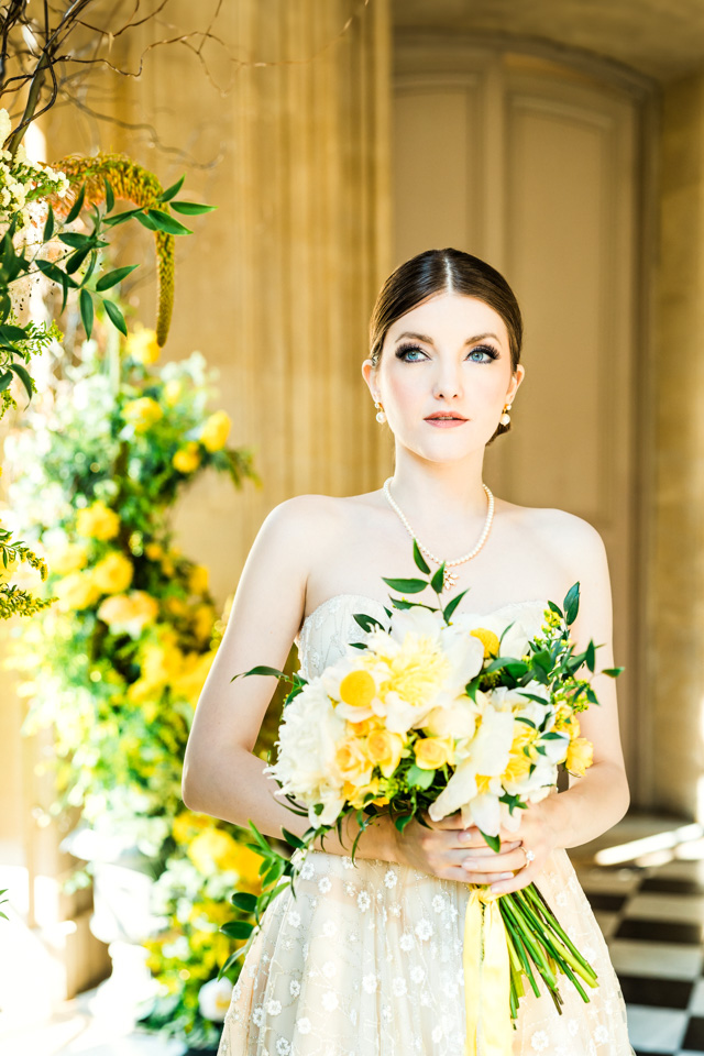 Die Braut hält beim Bridal Shooting ihren Brautstrauß fest. Sie schaut in die Ferne und befindet sich in einem Raum voller gelber Blumen.