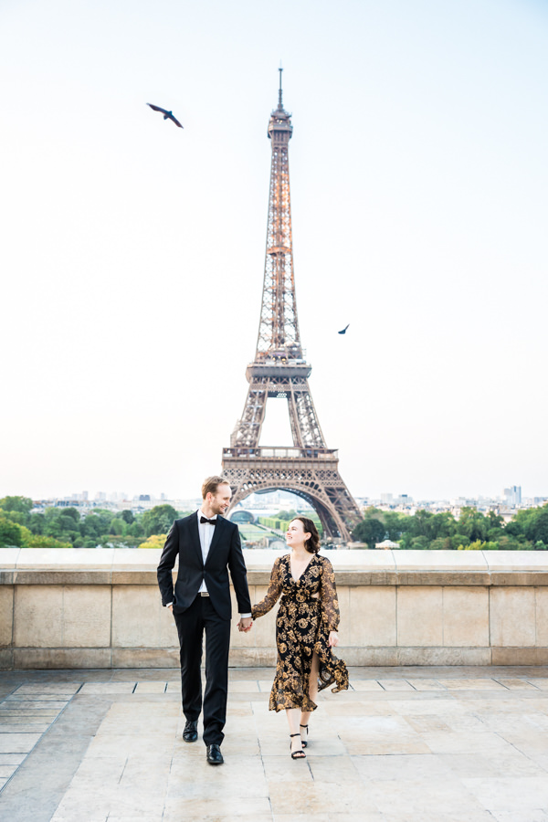 Romantisches Verlobungsshooting vor dem Eiffelturm in Paris. Beide gehen Hand in Hand und schauen sich dabei an. Tauben fliegen über ihren Köpfen durch die Luft.