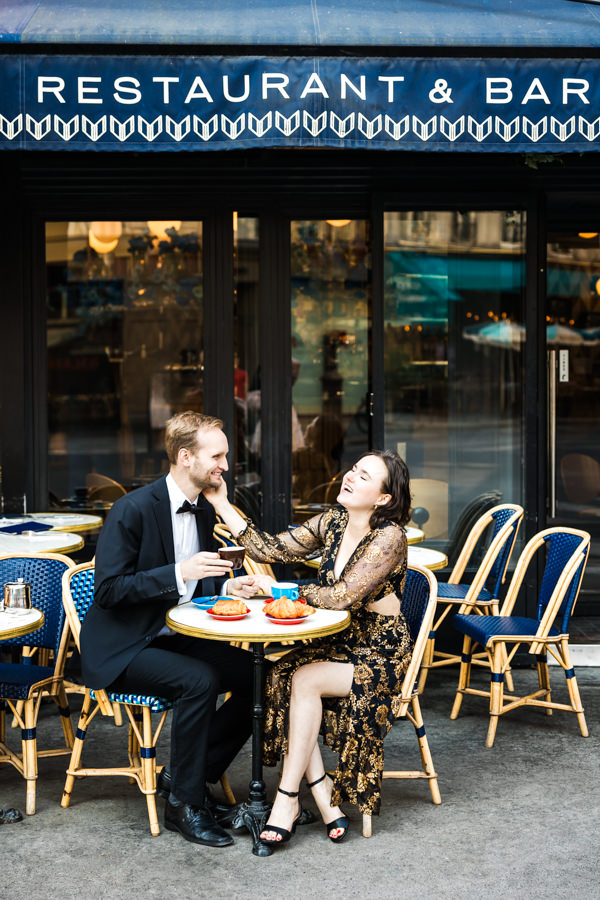 Paar sitzt beim Verlobungsshooting in einem romantischen Café. Sie streichelt sein Gesicht und beide lachen.