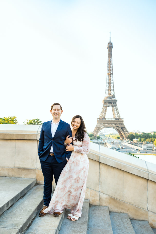 Pärchen steht beim Paarshooting in Paris auf einer Treppe vor dem Eiffelturm und ist glücklich.