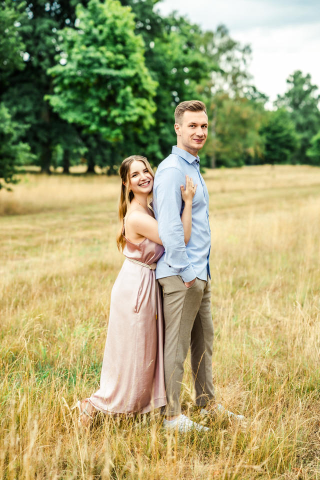 Paar steht beim Shooting im Georgengarten Hannover in einem weiten Feld. Im Hintergrund sind Bäume zu sehen.
